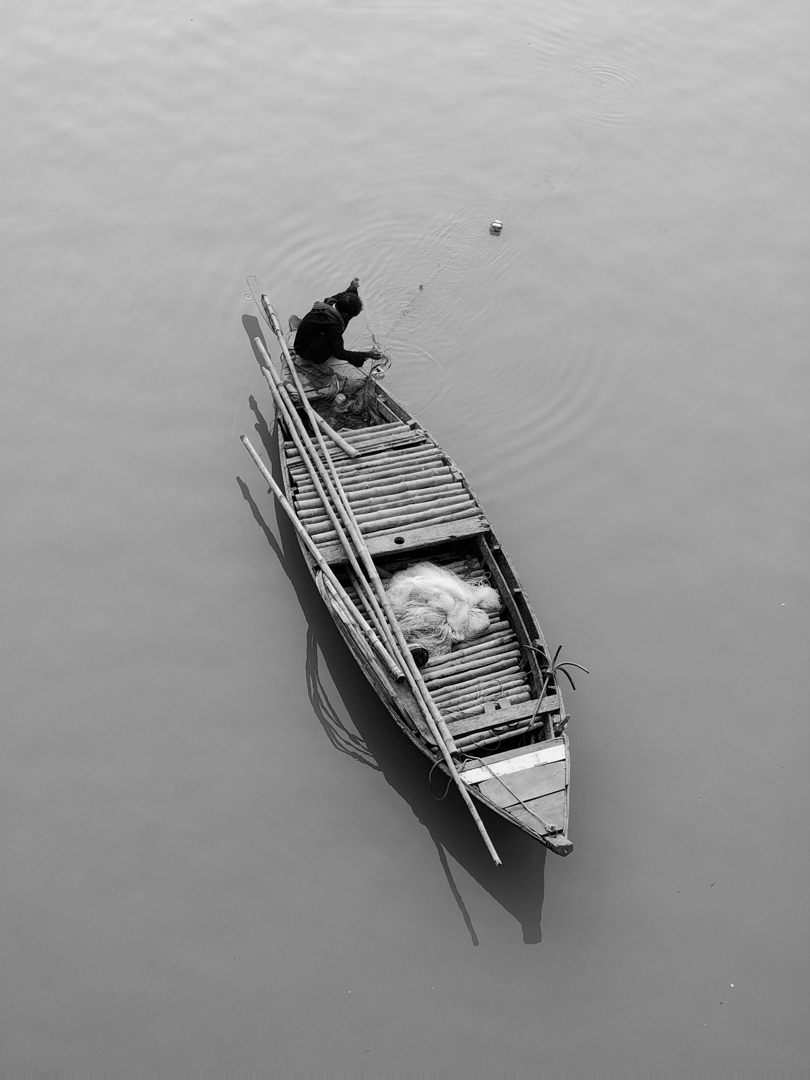 An aerial black and white shot of a solitary fisherman in a wooden boat on calm waters.