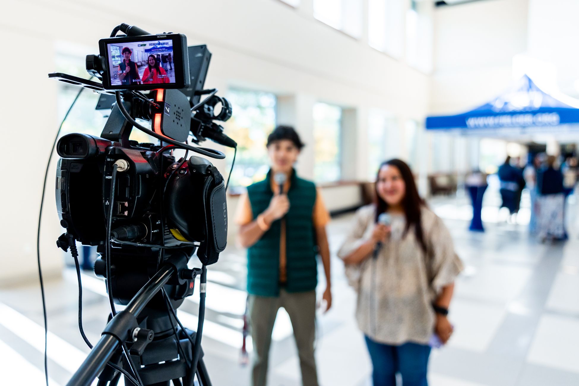 A professional video camera captures two interviewees in an indoor setting.