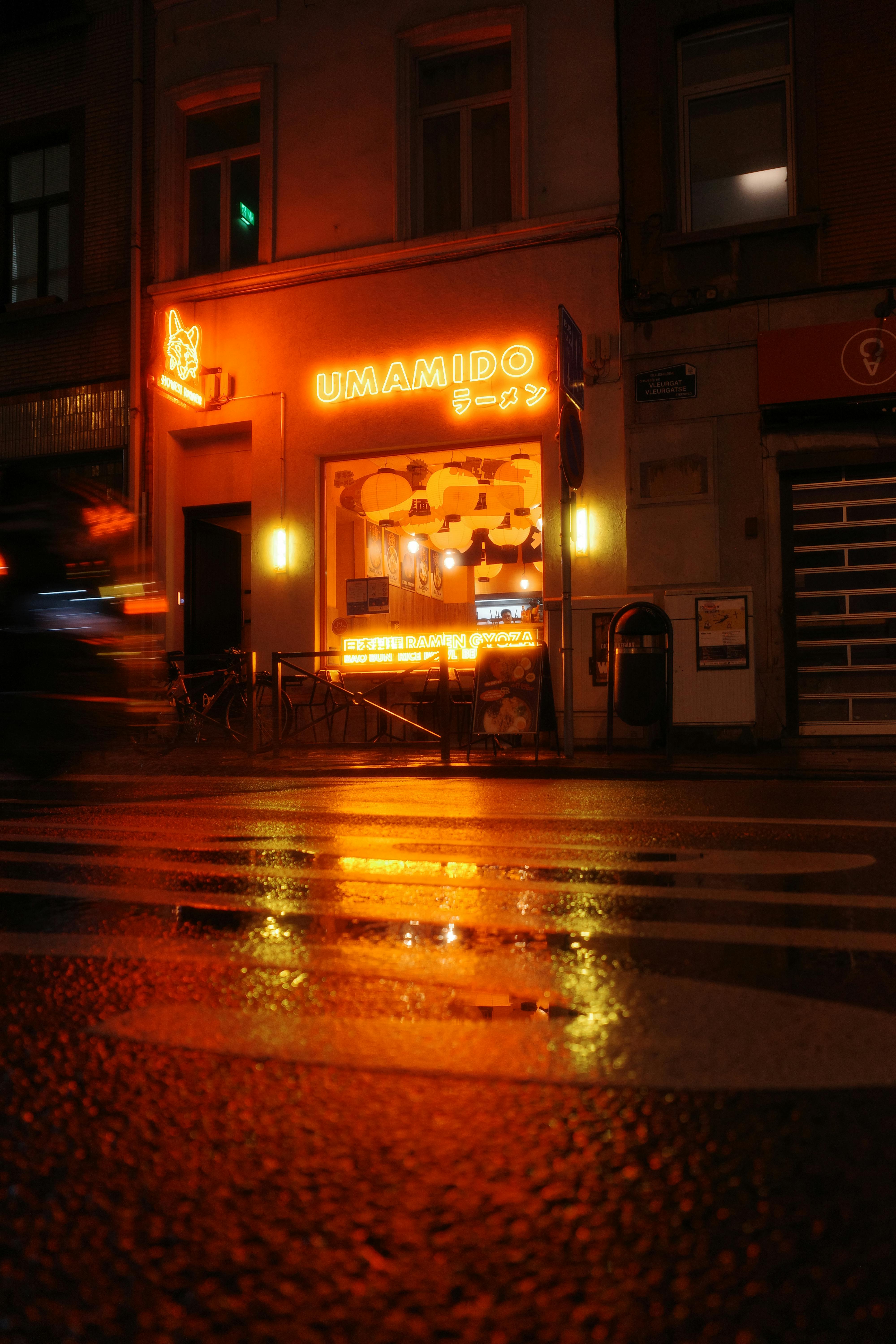 Vibrant neon lights illuminate Umamido restaurant in Brussels, reflecting on wet streets at night.