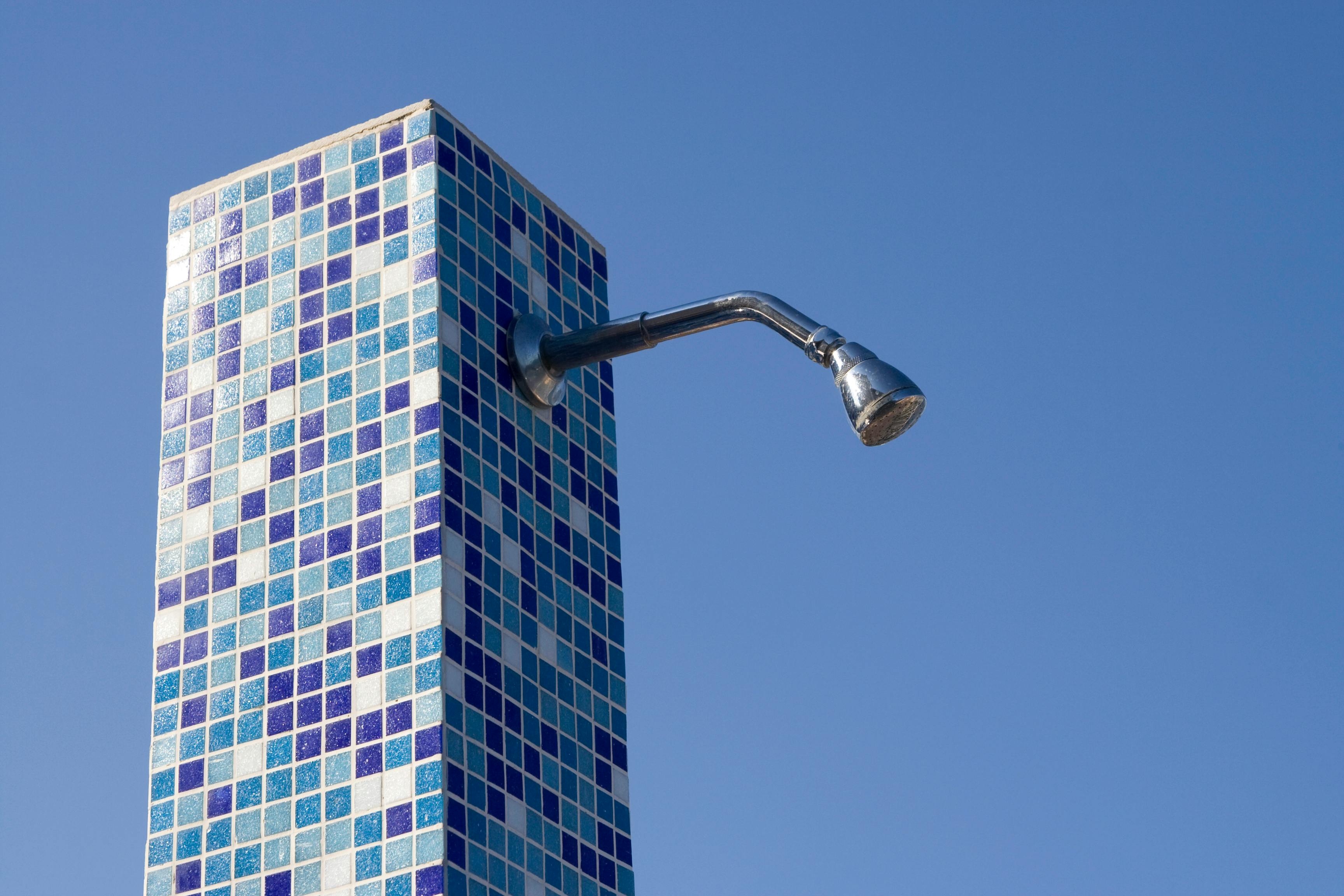 A modern outdoor shower with blue mosaic tiles and a chrome shower head under a clear blue sky.