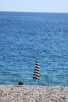 A lone striped umbrella stands on a pebble beach with the blue ocean in the background.