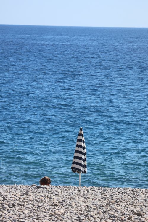 Free A lone striped umbrella stands on a pebble beach with the blue ocean in the background. Stock Photo