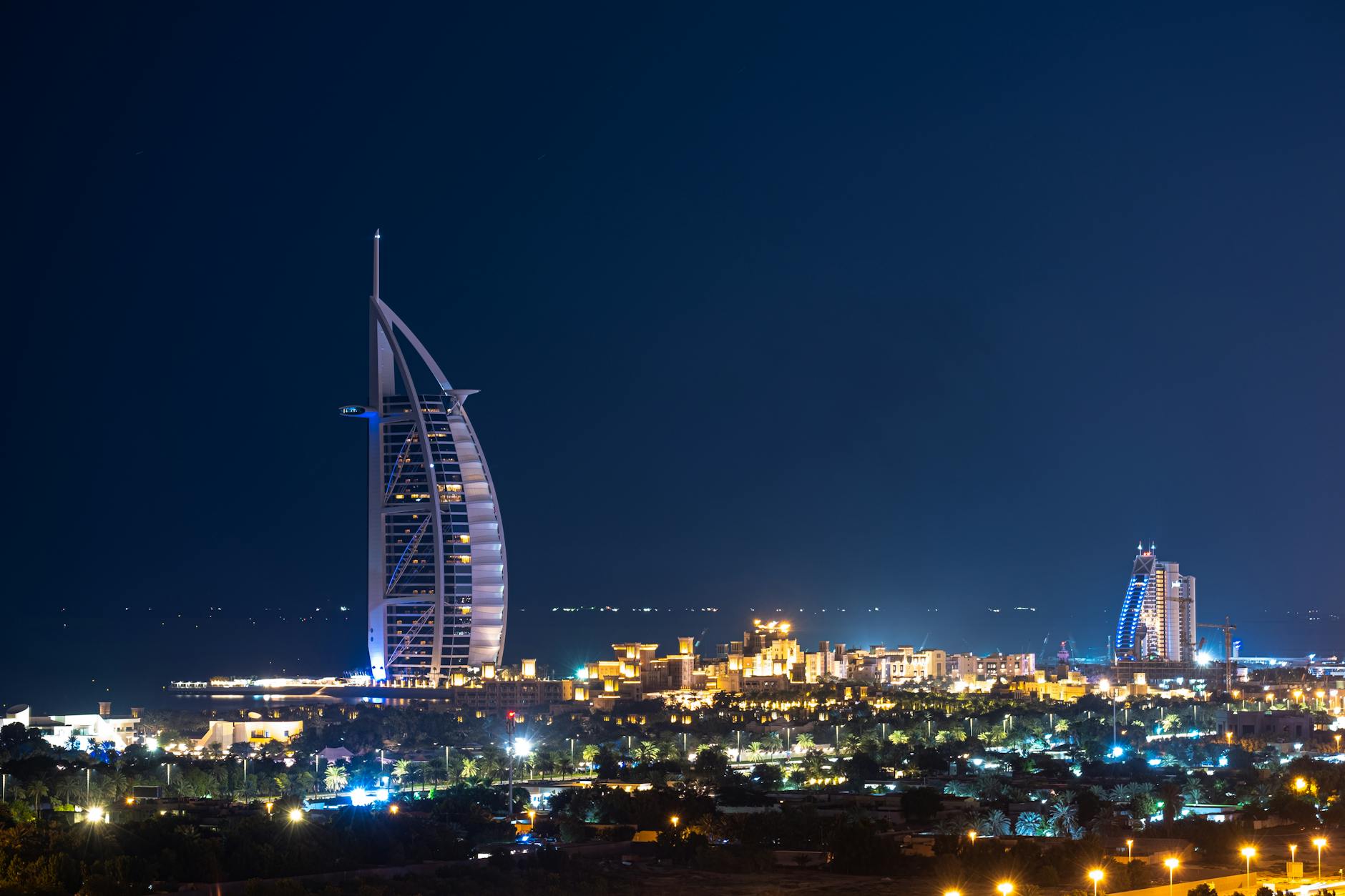 Dark blue cloudless sky above tall Burj Al Arab skyscraper hotel surrounded by buildings glowing in dark