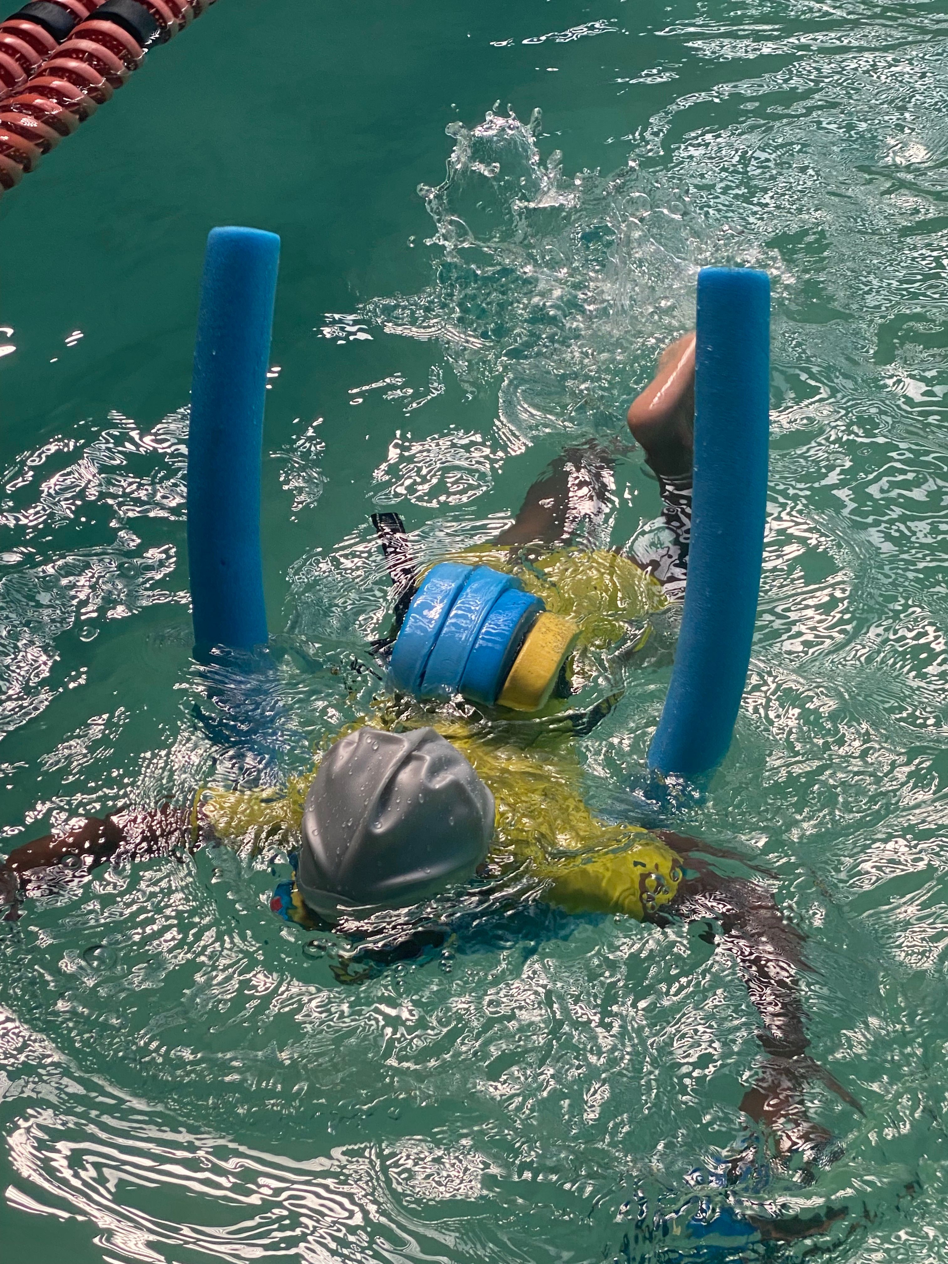 A child practicing swimming with pool noodles and a floatation belt in an indoor pool.