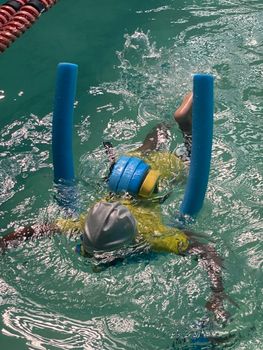 A child practicing swimming with pool noodles and a floatation belt in an indoor pool.