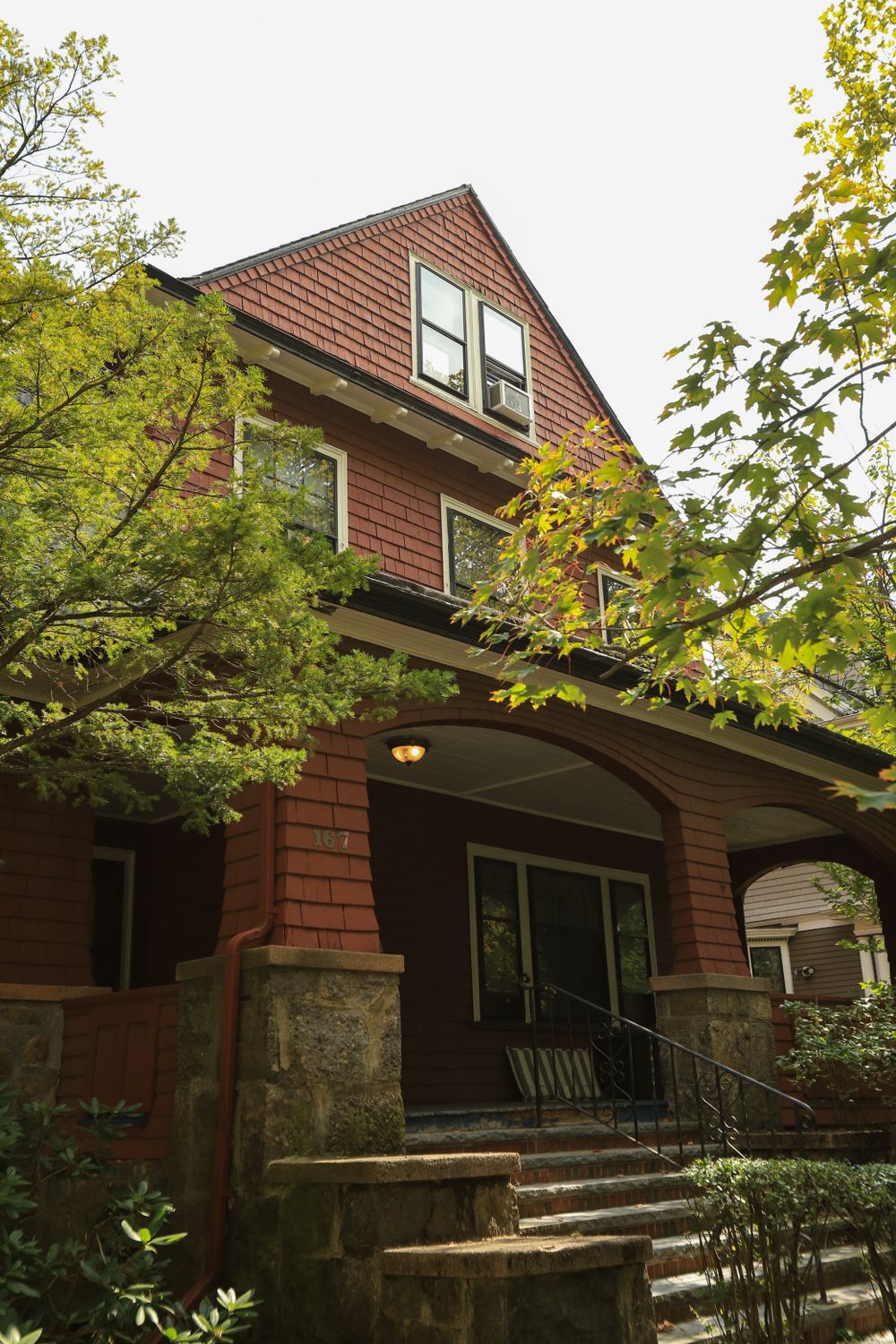 A shingled foursquare house with a deep stone porch, half hidden behind maple trees