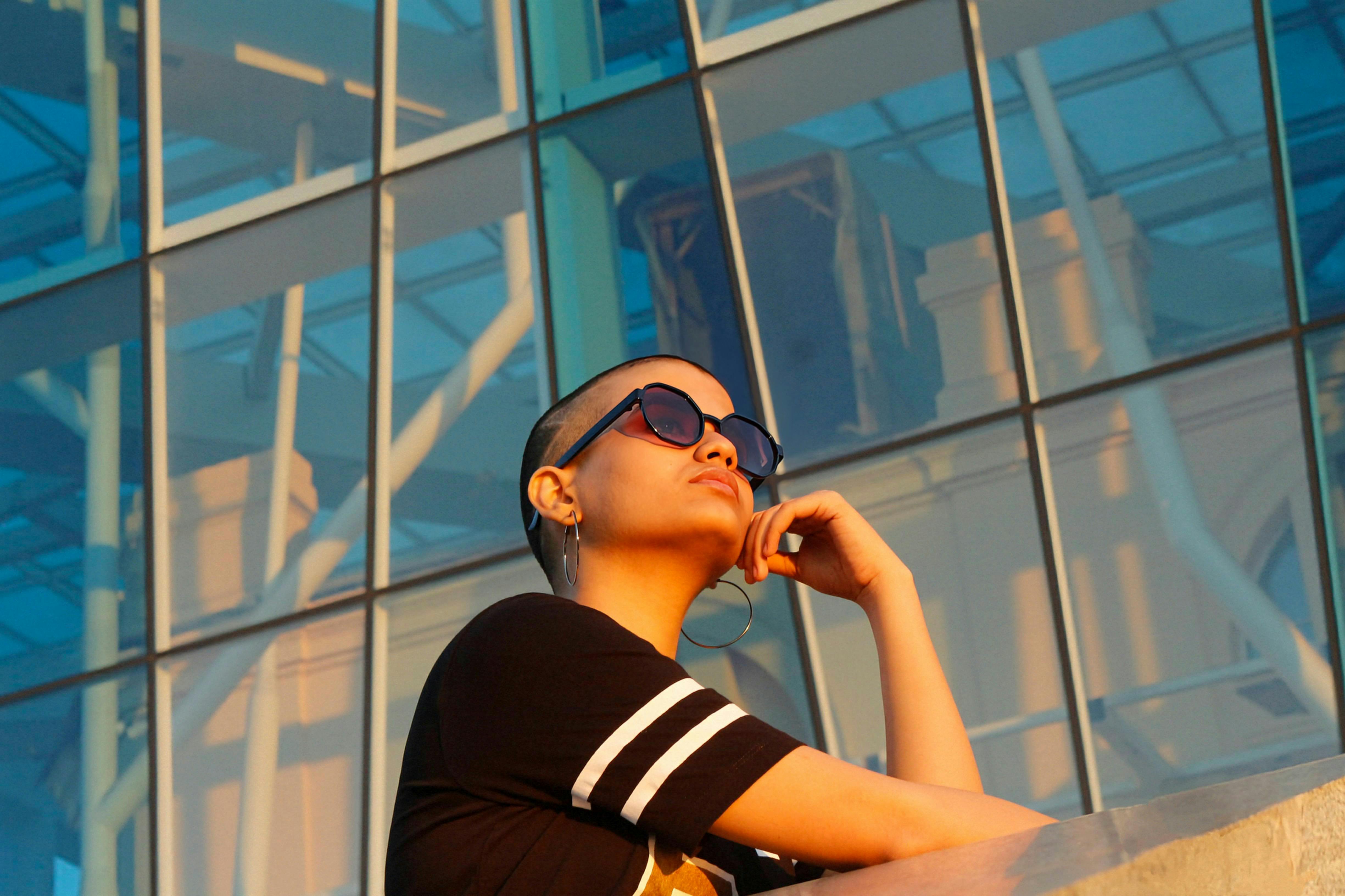 무료 선글라스를 낀 세련된 여성이 일몰 무렵 현대적인 유리 건물을 생각에 잠긴 듯 바라보고 있습니다. 스톡 사진