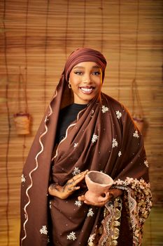 Smiling African woman in traditional attire holding a clay cup indoors.