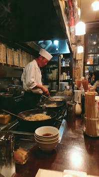 Japanese chef preparing noodles in a cozy, dimly-lit restaurant kitchen.