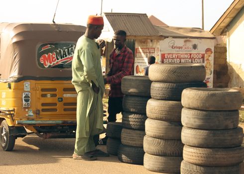 Two men conversing near a stacked tire shop with a yellow tuk-tuk in a bustling street setting.