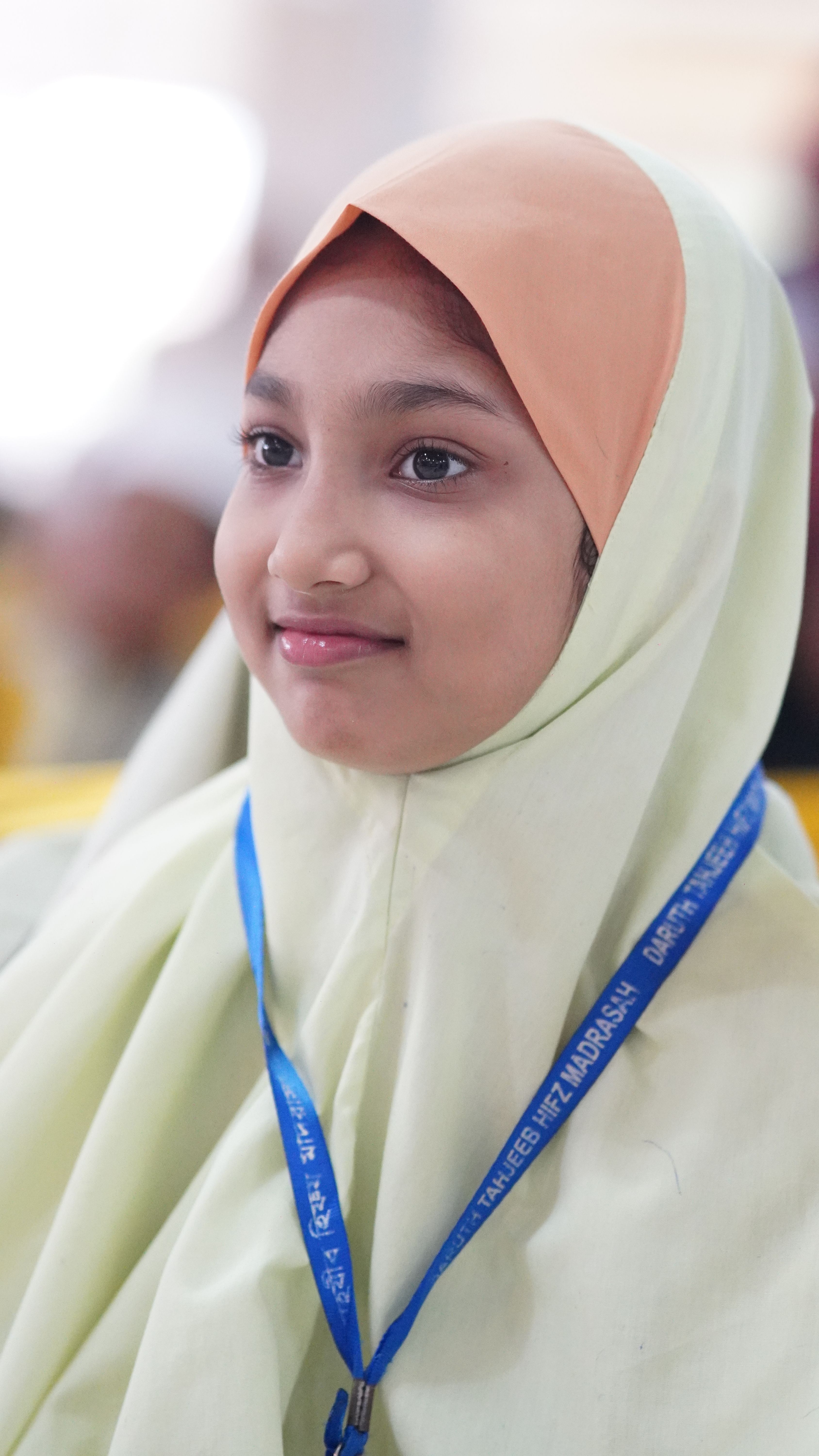 Close-up of a young girl wearing hijab and lanyard, smiling in a bright setting.