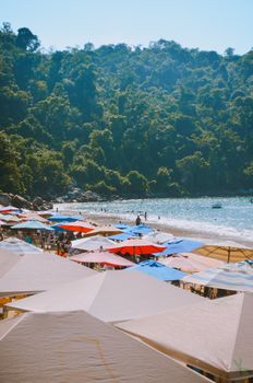 A vibrant beach with colorful umbrellas under a bright sunny sky, set against lush green hills.