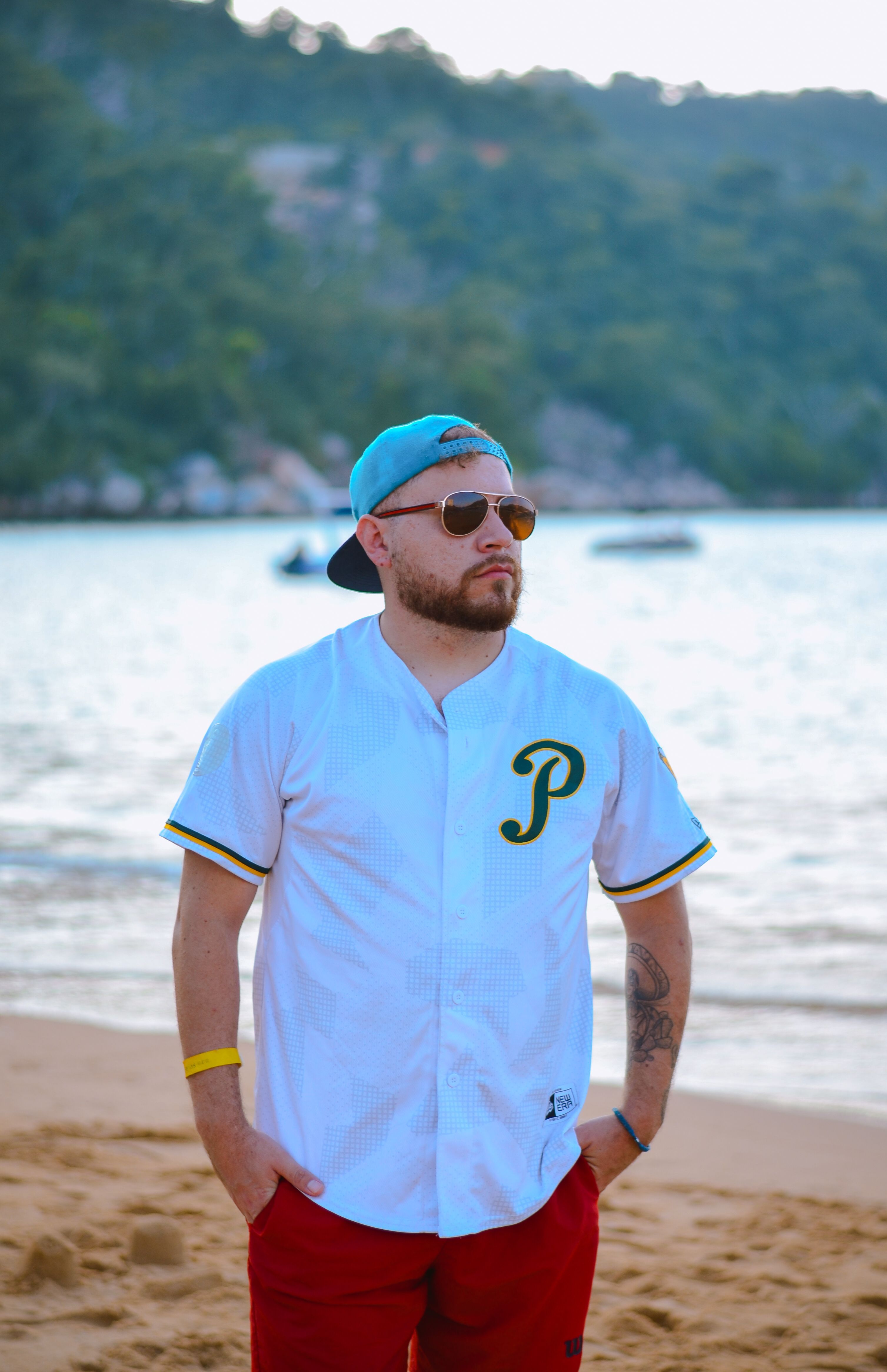 Man wearing a baseball shirt and cap standing on a beach with a scenic background.