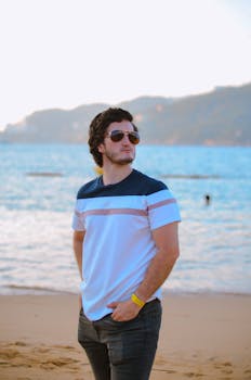 Young man in casual wear stands confidently on a sunny beach with ocean backdrop.