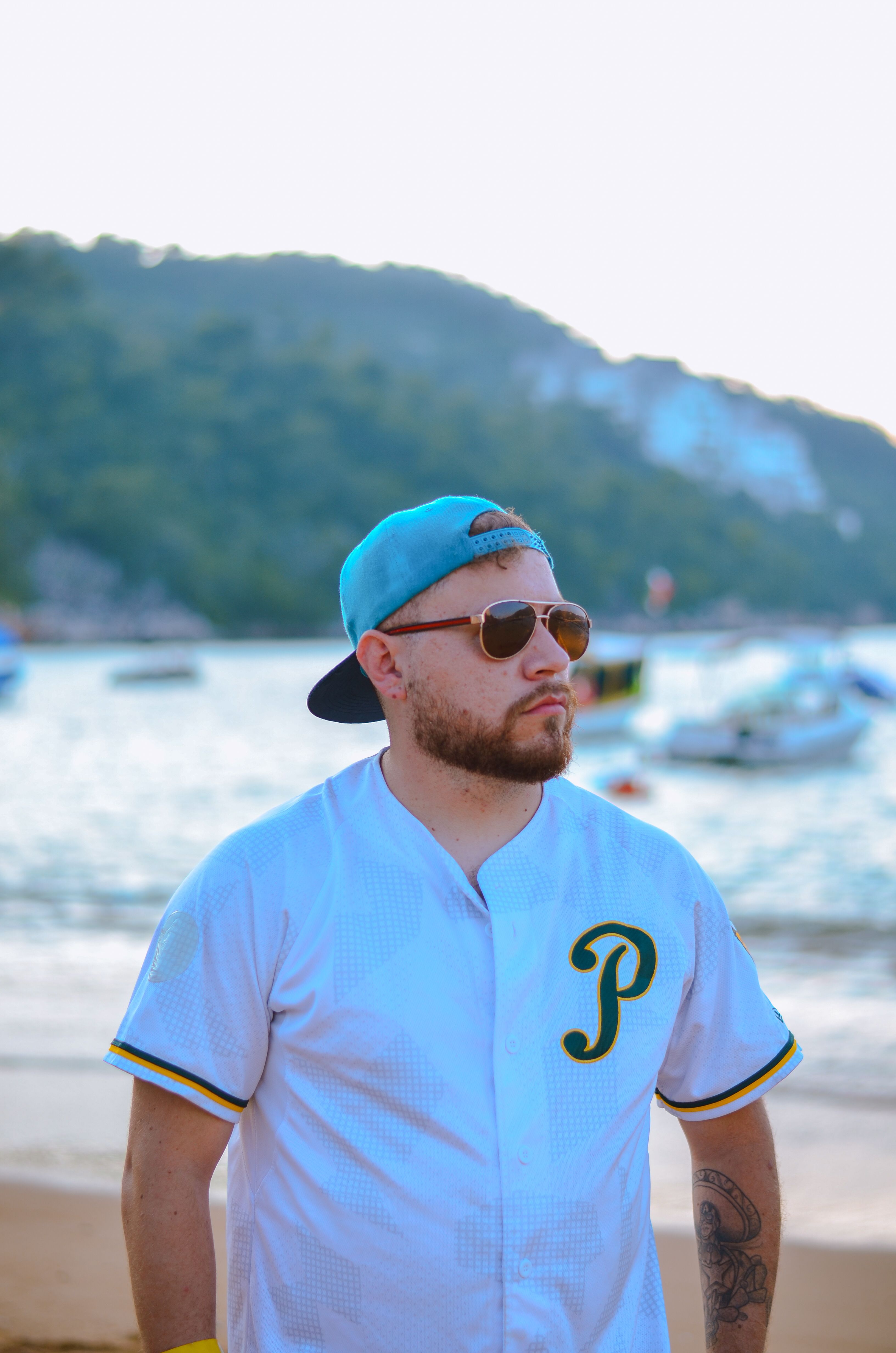 Man in sunglasses and cap enjoying a sunny day by the seaside with boats in the background.