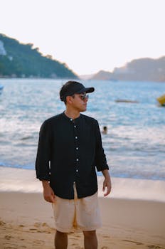 A young man in casual attire stands on a scenic beach during sunset, exuding tranquility.