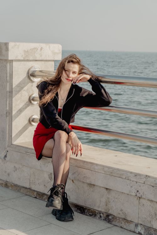 Free A fashionable woman in red skirt sitting by a seaside railing, exuding confidence. Stock Photo