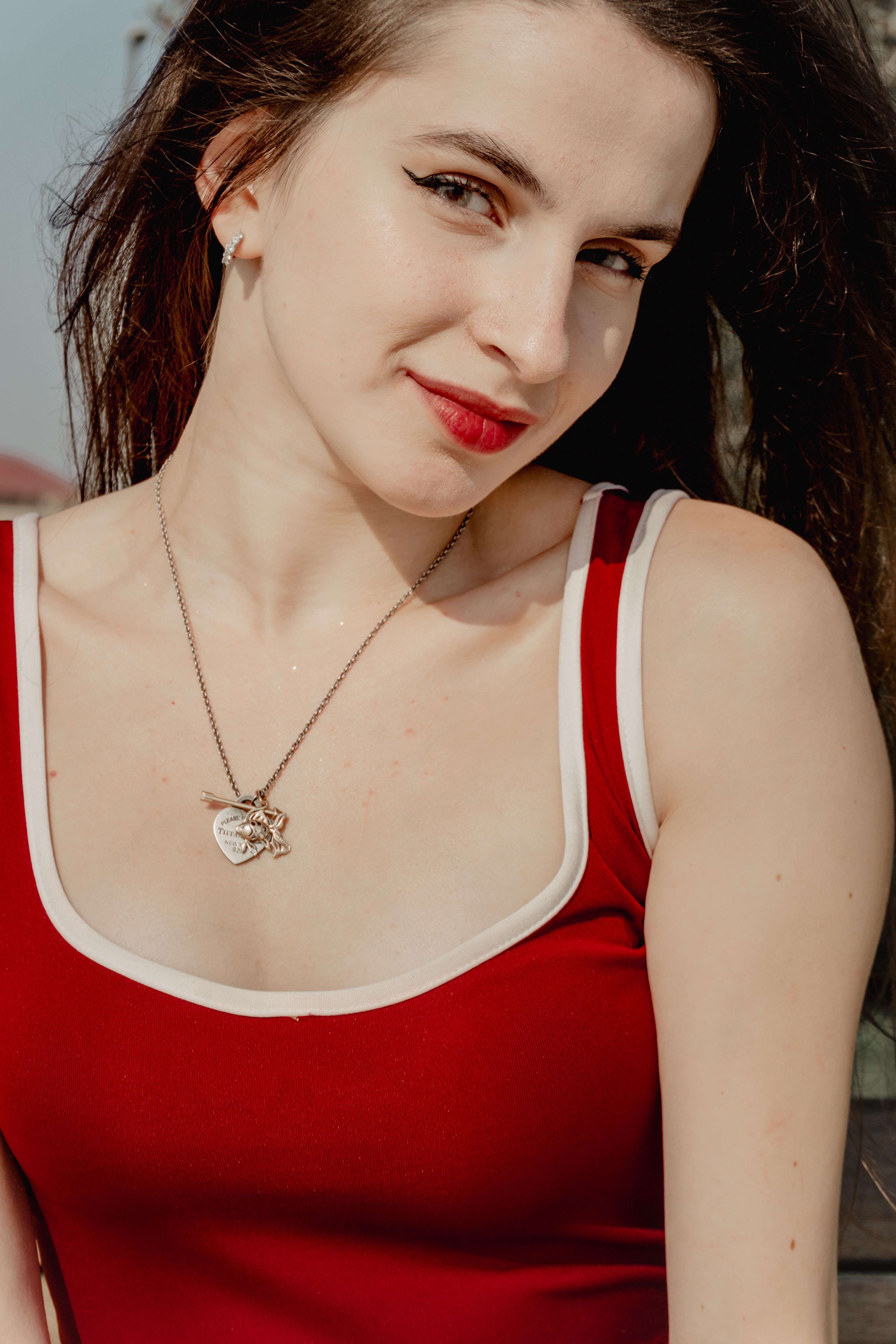 Portrait of a young woman wearing a red tank top and silver necklace enjoying sunlight outdoors.
