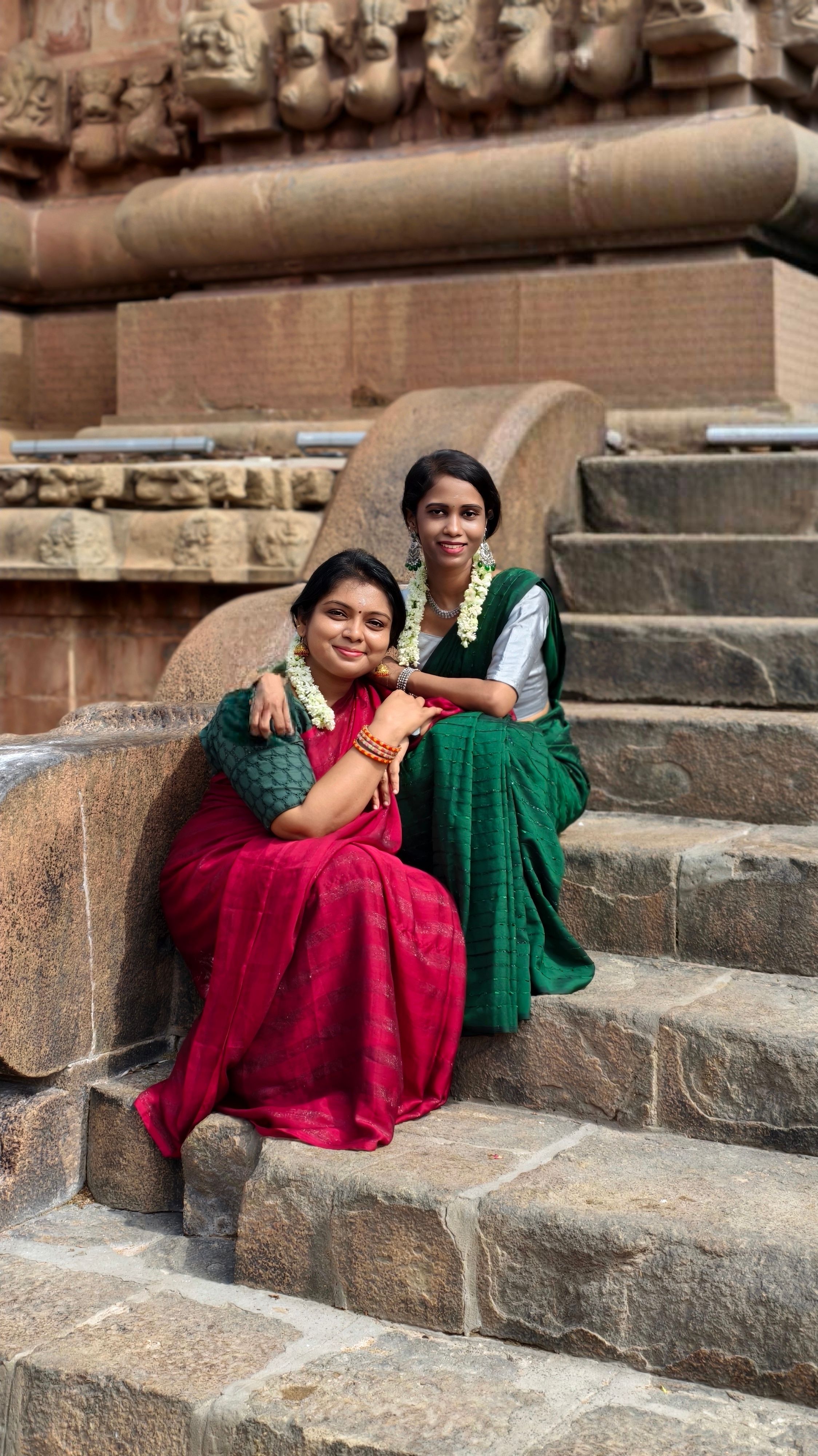 Two women in elegant sarees pose on stone steps at Thanjavur Temple, India.