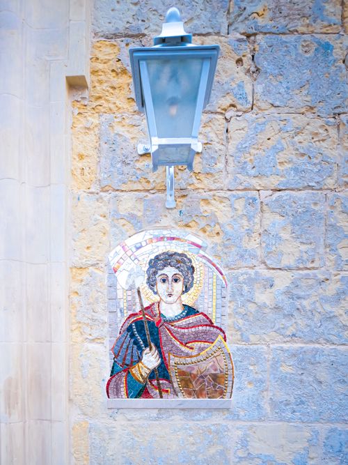 Free A colorful mosaic depicting a saint on a stone wall with a street lamp in Malta. Stock Photo