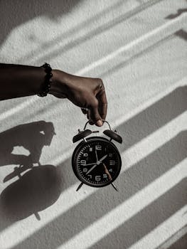 Close-up of a hand holding a vintage alarm clock casting shadows in daylight.