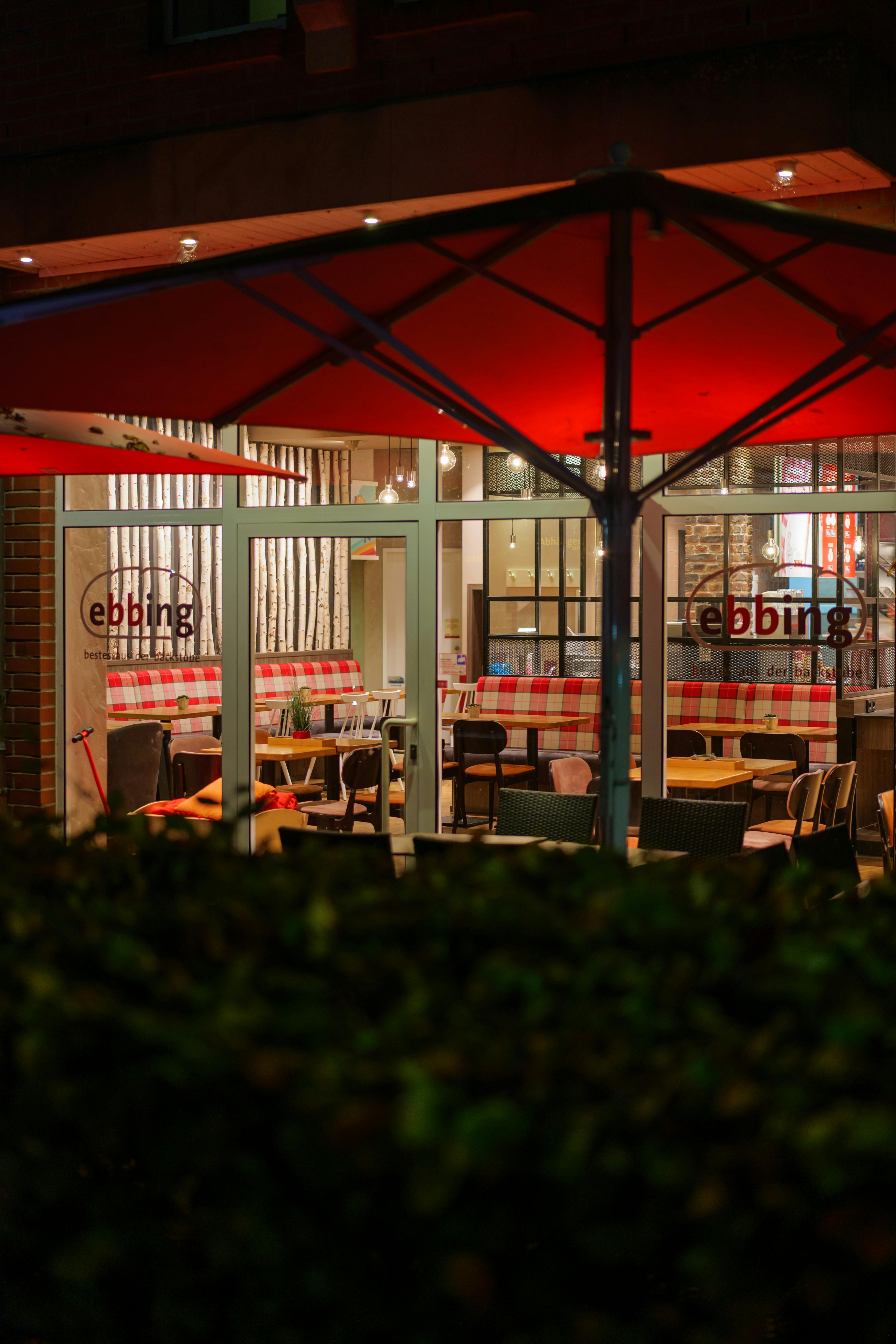 A charming view of a cozy café with red umbrellas, seen at night through large windows.
