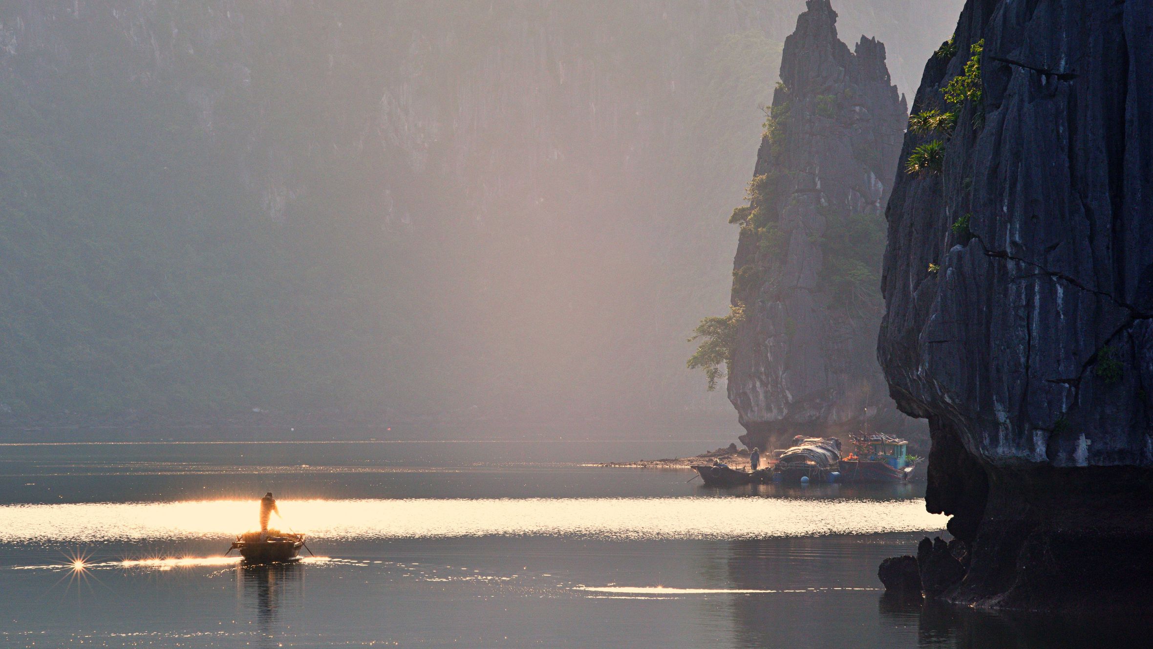 Morning light shimmers on Halong Bay as a lone fisherman navigates tranquil waters.