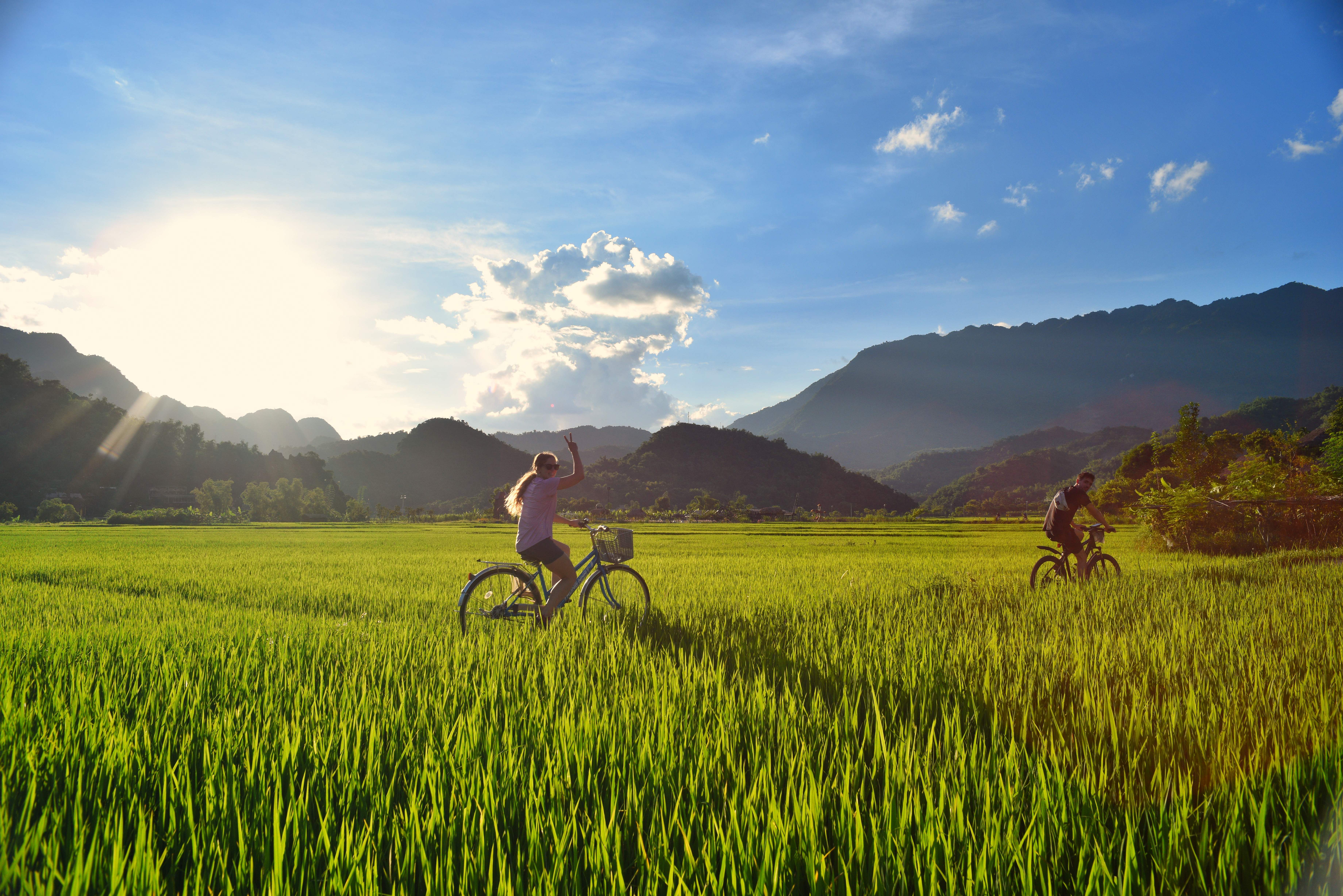 Two cyclists riding through lush green fields with mountain backdrop under a bright sunny sky.