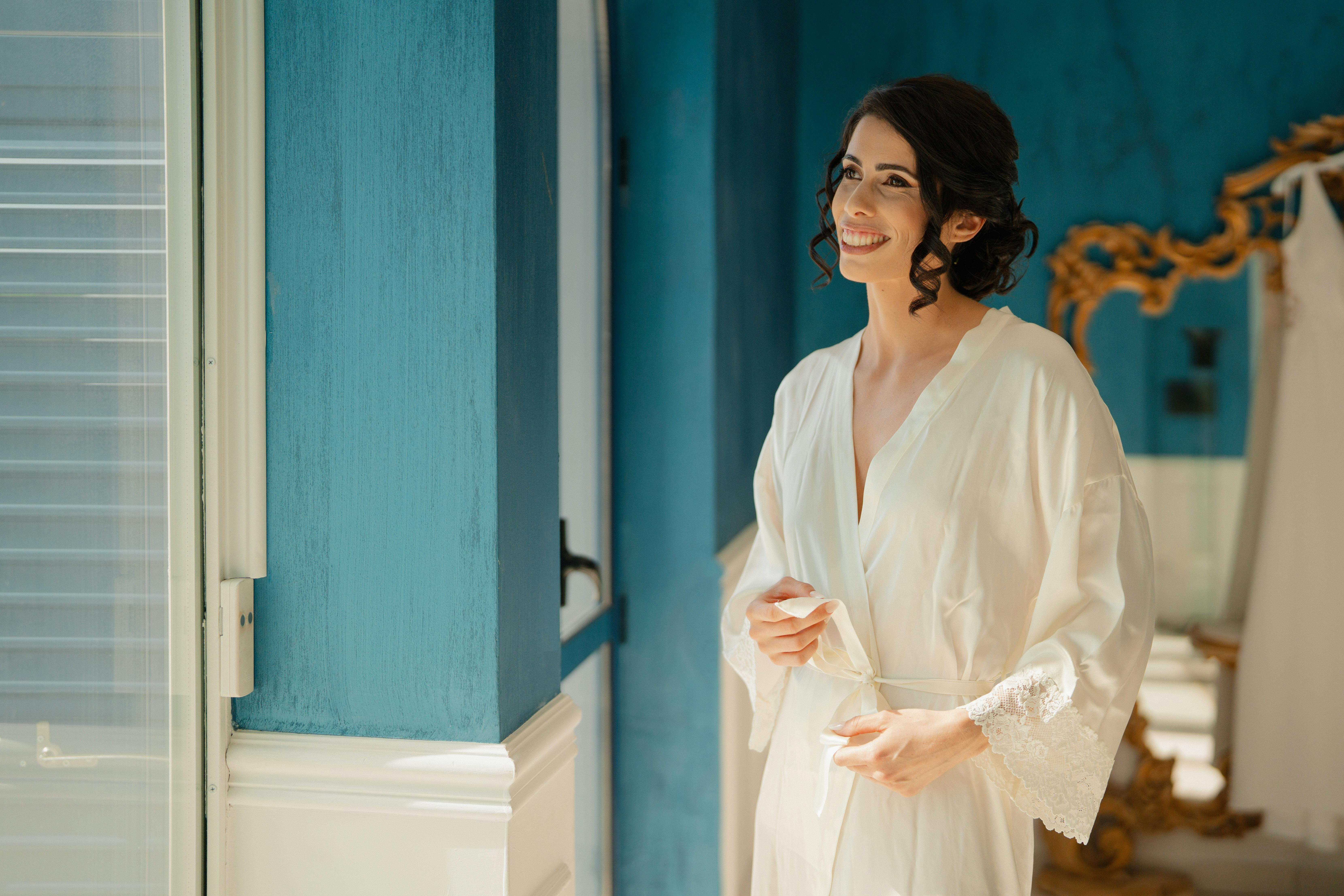 A joyful bride in a white robe smiling indoors with natural light streaming through a window.