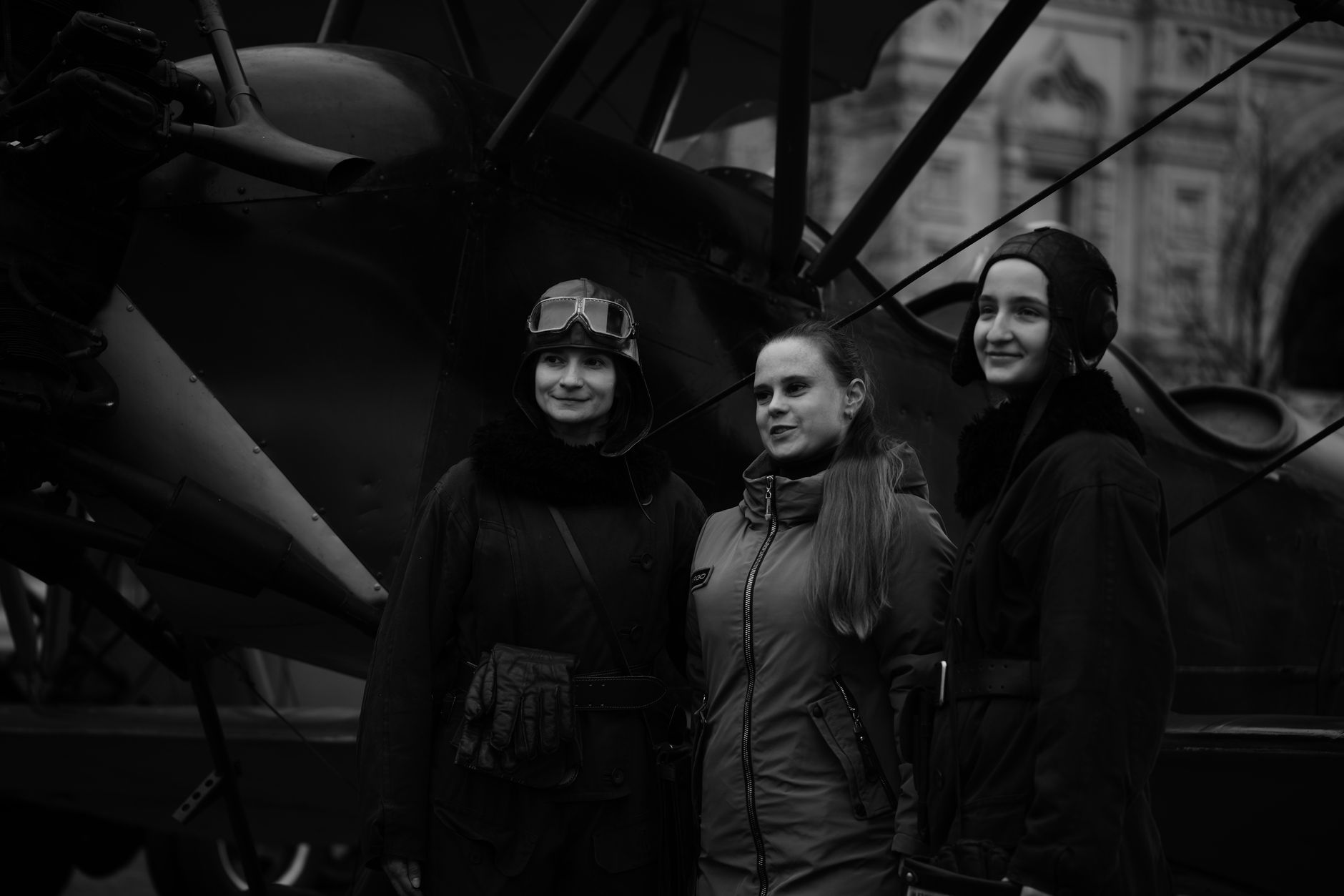 Three women in vintage aviator attire pose in front of a classic aircraft, capturing a nostalgic aviation theme.