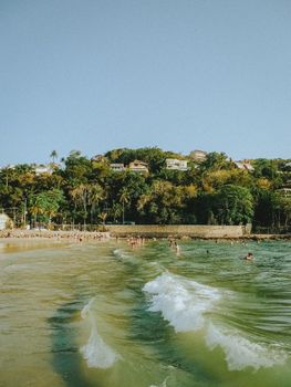 Scenic view of a tropical beach with waves and hillside houses under clear skies.