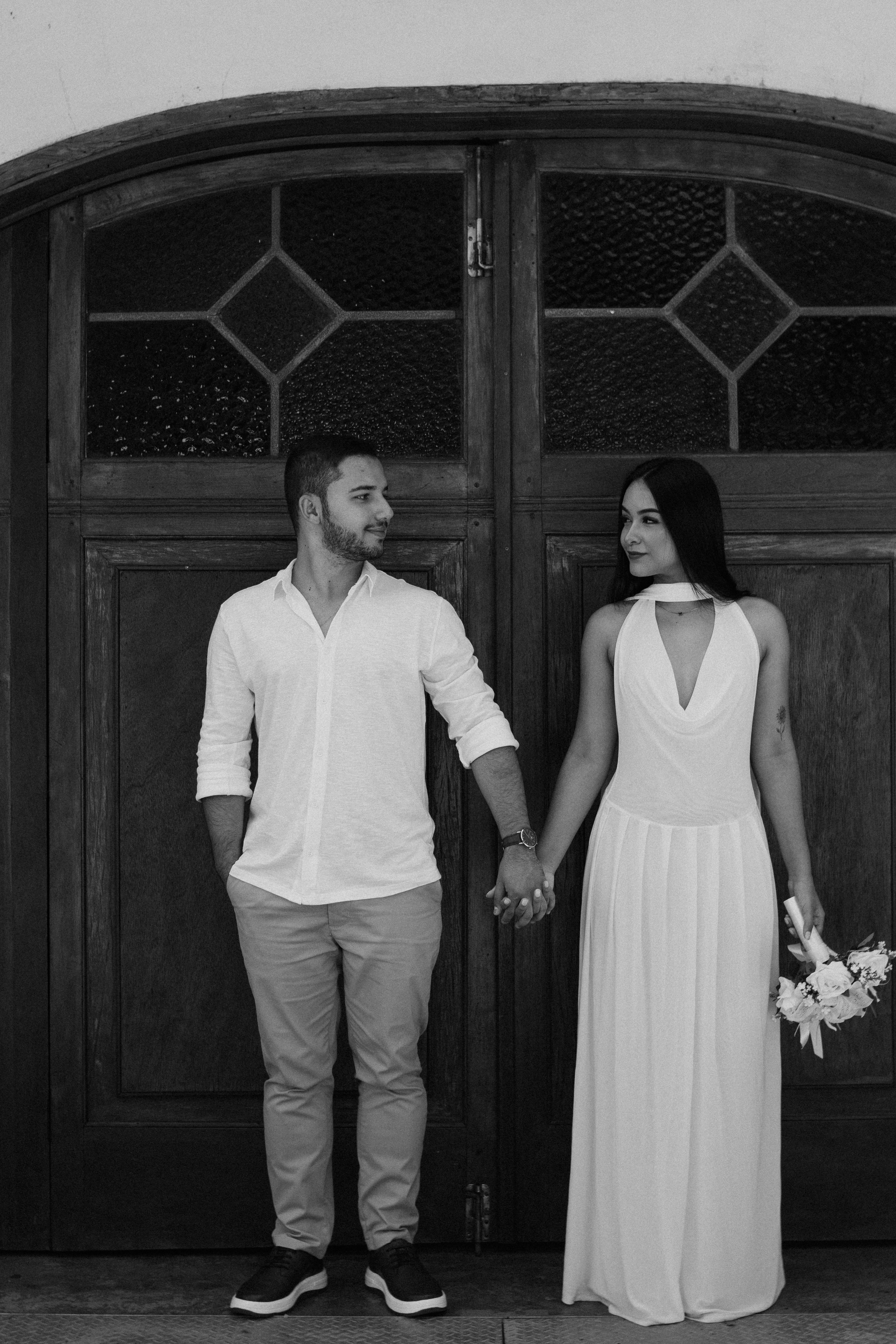 Elegant couple standing together, holding hands in front of a wooden door, black and white photo.