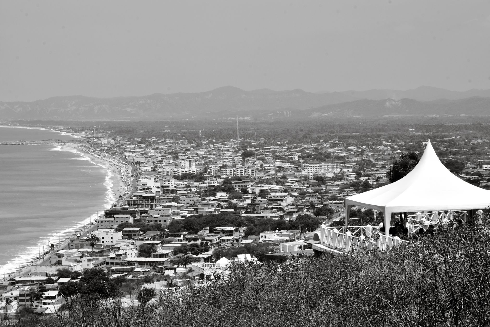 Black and white aerial view of a coastal city in Ecuador with a beach and mountains in the background.