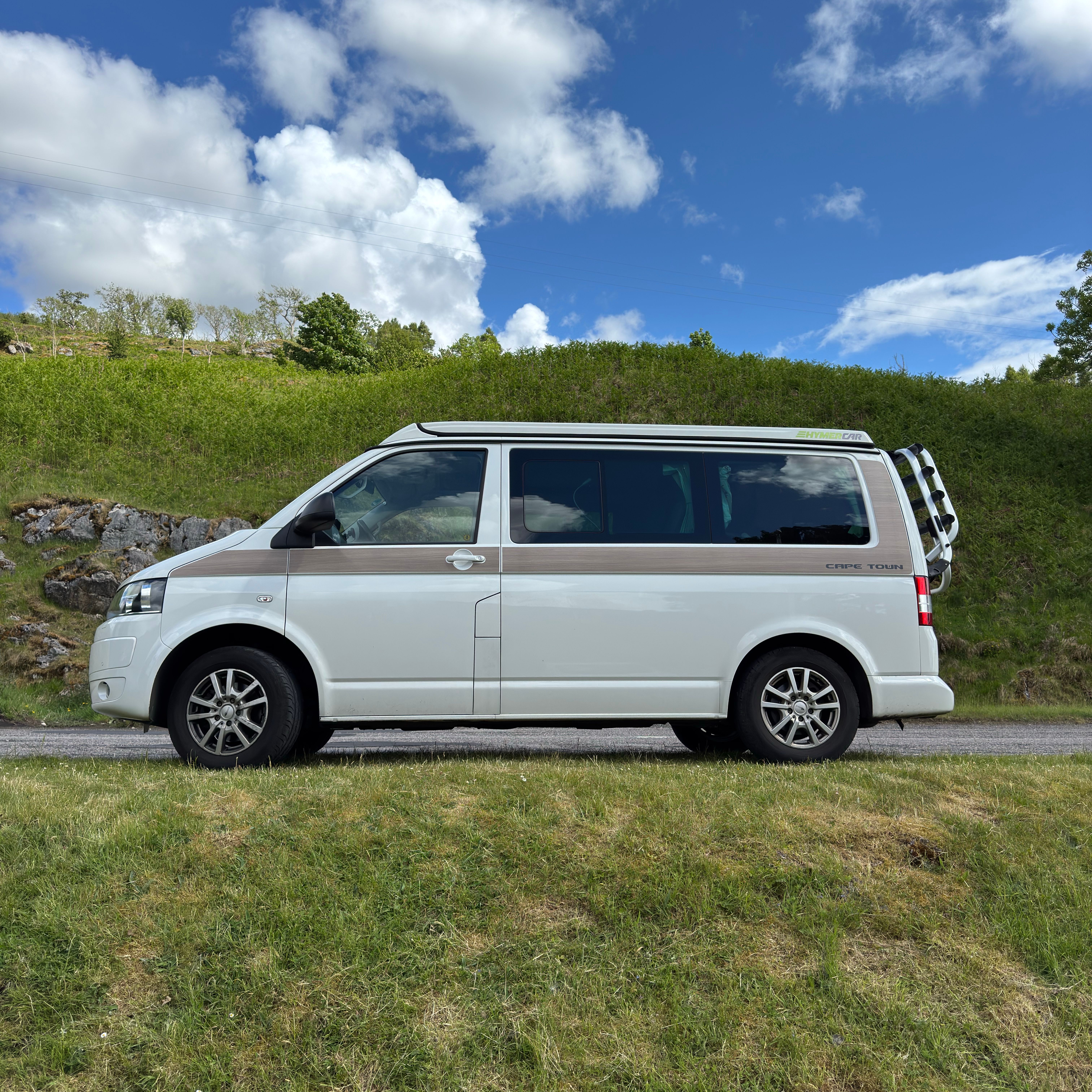 A white camper van parked on a country road amidst lush greenery and blue skies.