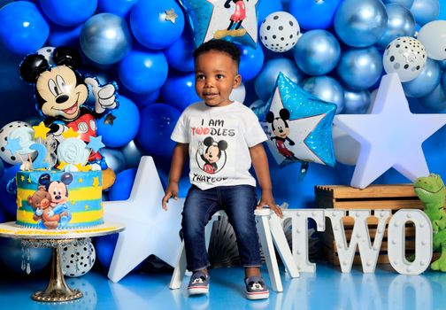 Young child celebrating a colorful Mickey Mouse themed birthday party with balloons and cake.