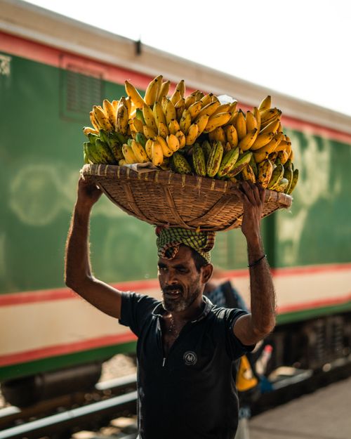 Free A man carries a basket of bananas on his head at a bustling Dhaka railway station. Stock Photo
