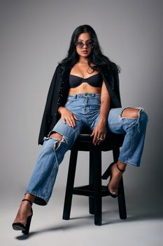 Confident woman in trendy outfit poses on stool with a modern studio backdrop.