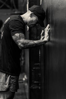 Black and white image of a tattooed man leaning against a wall, expressing contemplation.