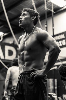 Black and white photo of a fit man resting in a gym, showcasing muscles and determination.