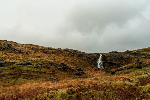 Free A scenic view of a small waterfall cascading down rocky hills surrounded by grass under a cloudy sky. Stock Photo