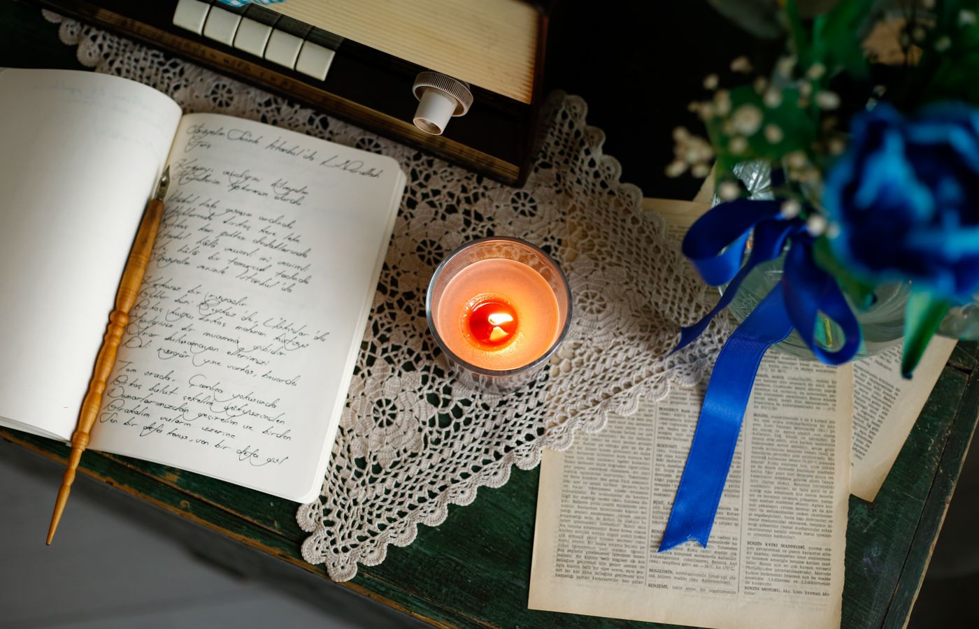 A cozy desk setting with a lit candle, open journal, lace doily, and blue floral bouquet.