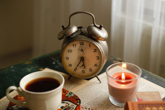 A cozy setting with a vintage alarm clock, coffee, and candle on a table.