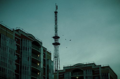 Free A cityscape featuring a communication tower rising above modern buildings at dusk with birds flying. Stock Photo