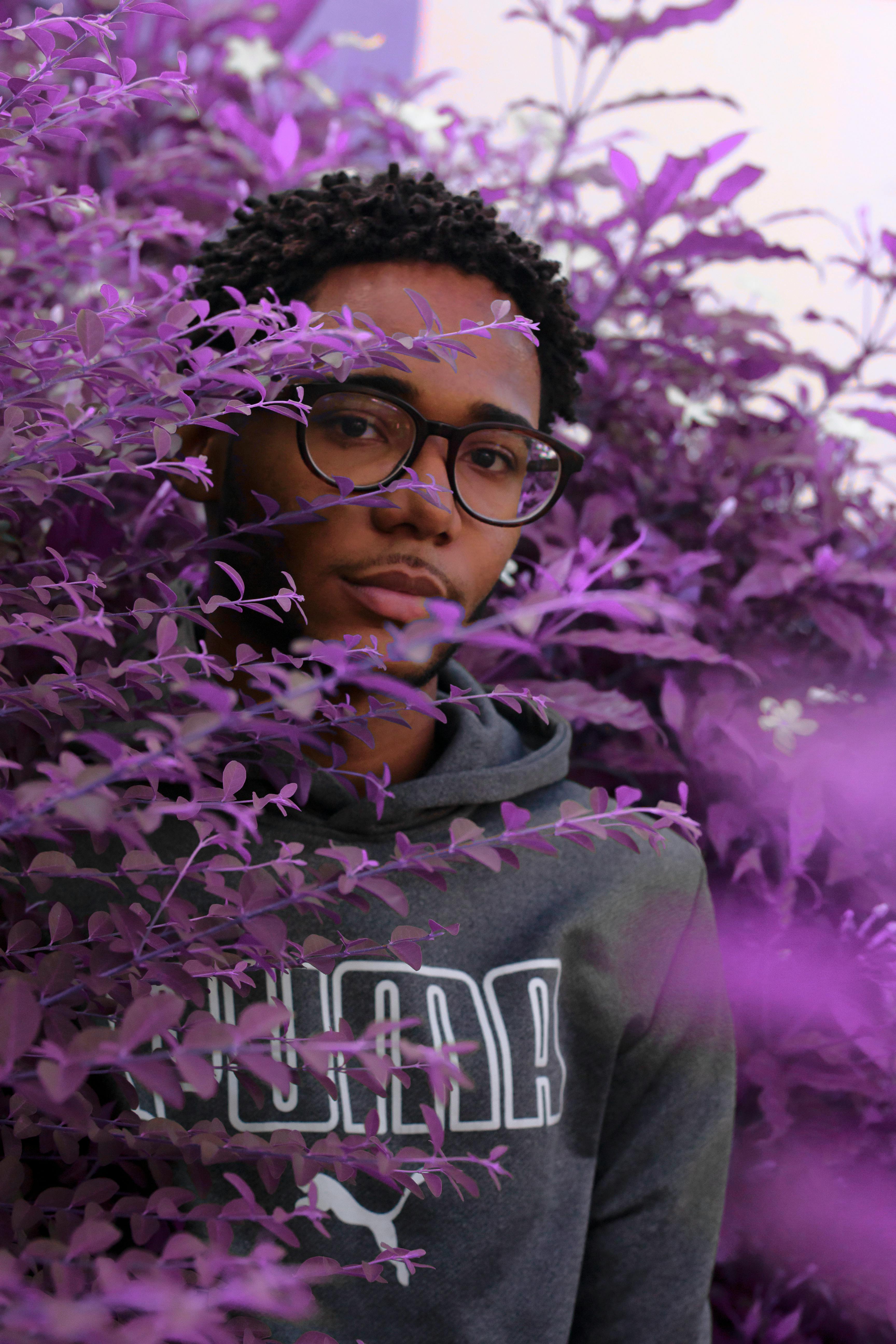 A fashionable young man in glasses posing surrounded by striking purple plants outdoors.