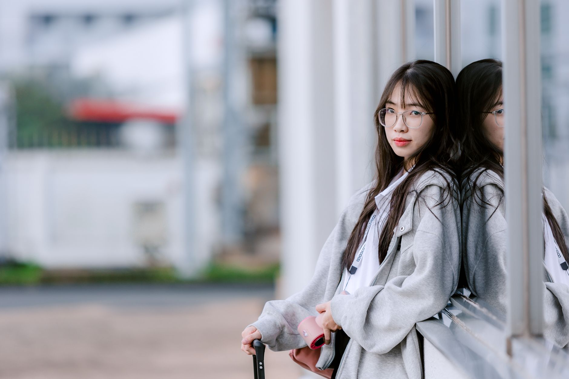 Stylish young woman in grey jacket leaning against a glass window, reflecting serene contemplation.