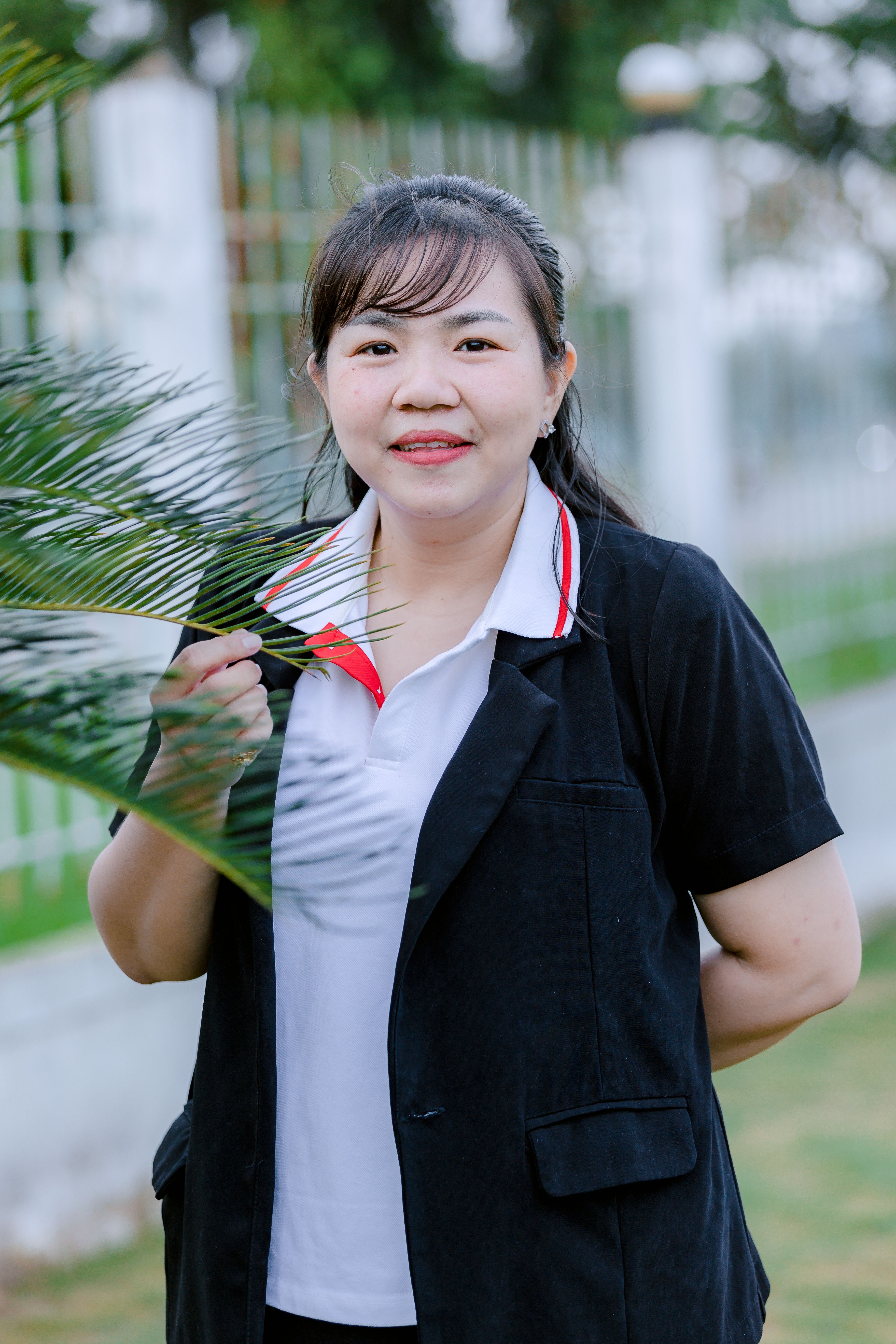 Young woman smiling and holding a palm leaf outdoors, dressed in casual attire.