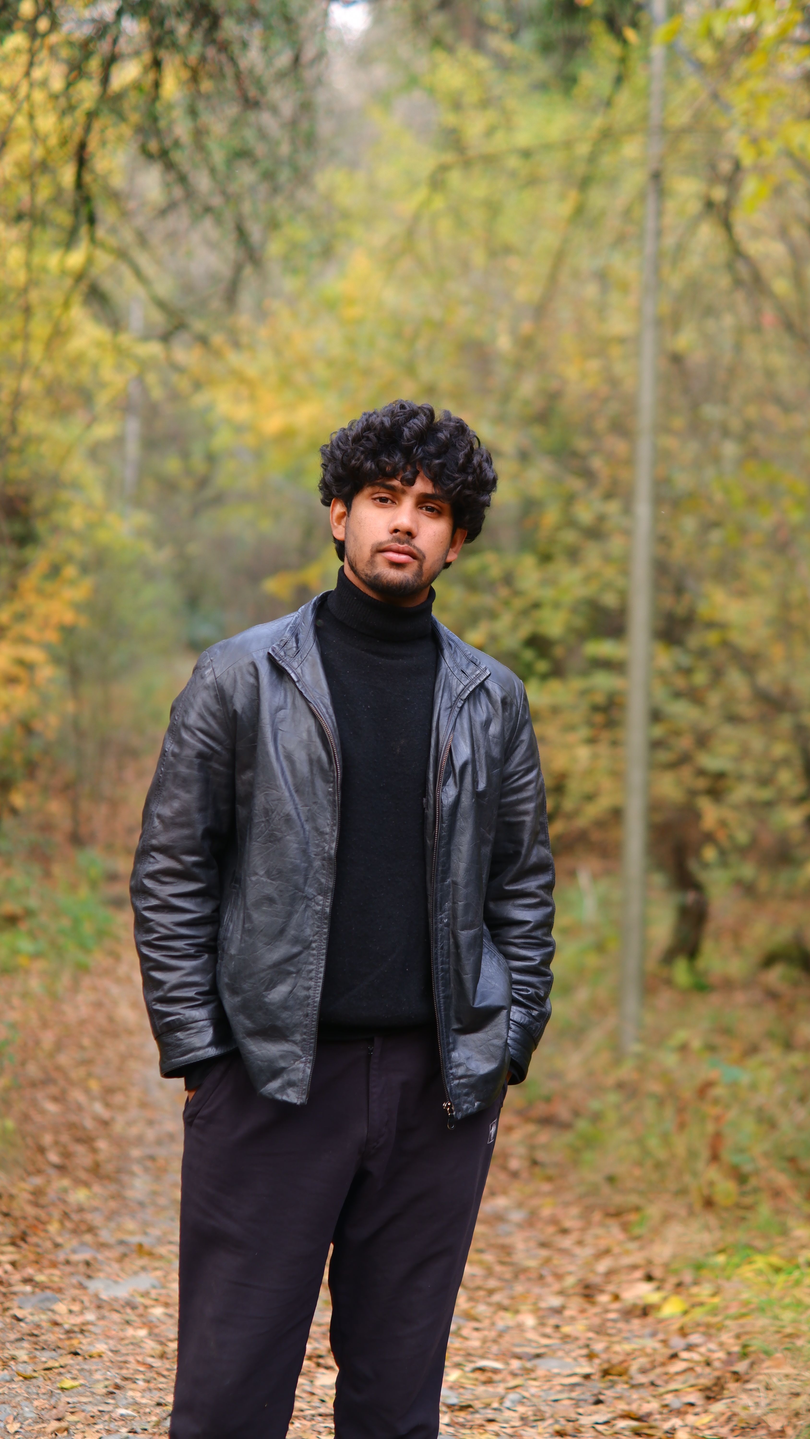 A man wearing a black leather jacket poses in a serene autumn forest setting.
