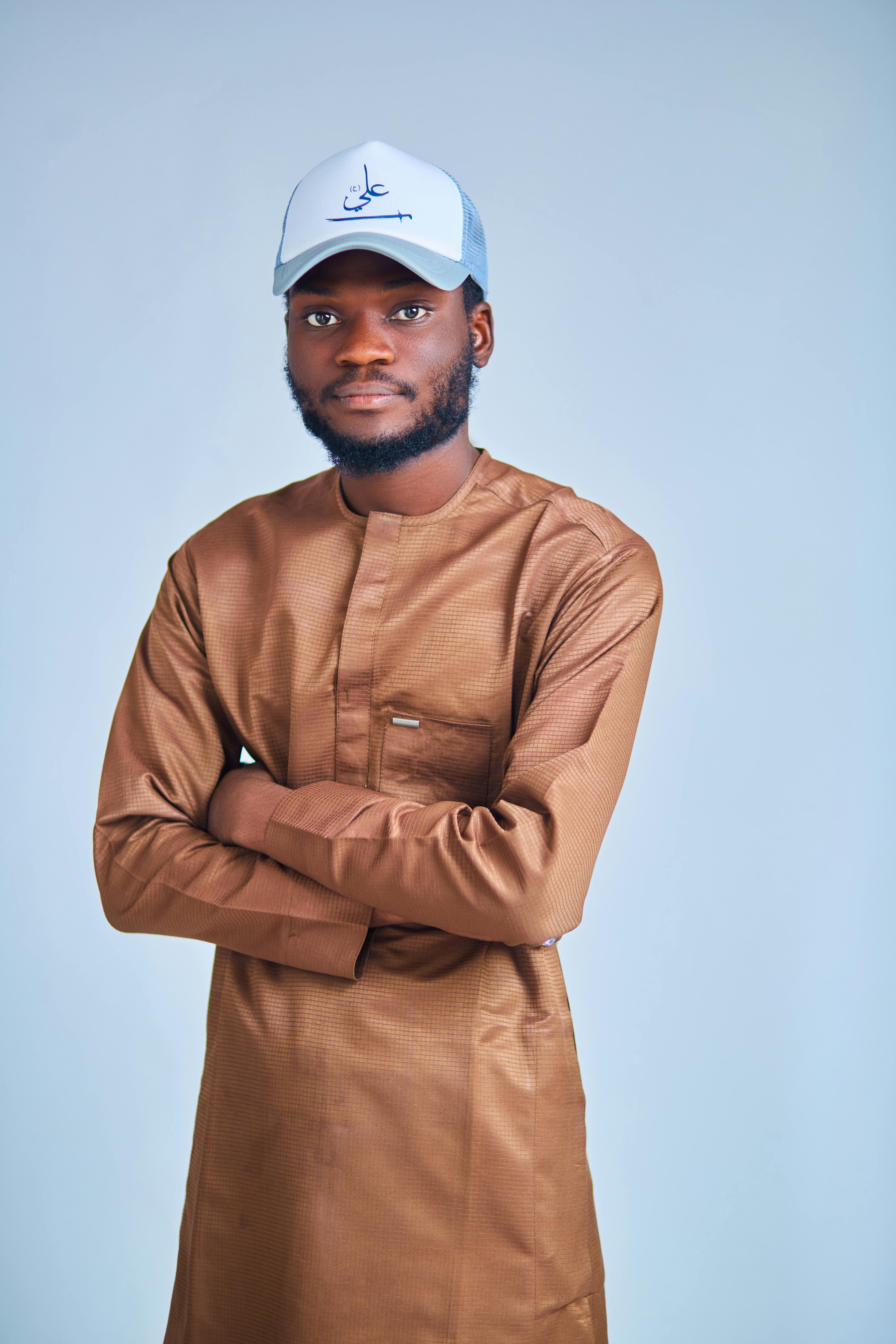 Confident man wearing traditional brown outfit and cap, posing indoors with arms crossed.
