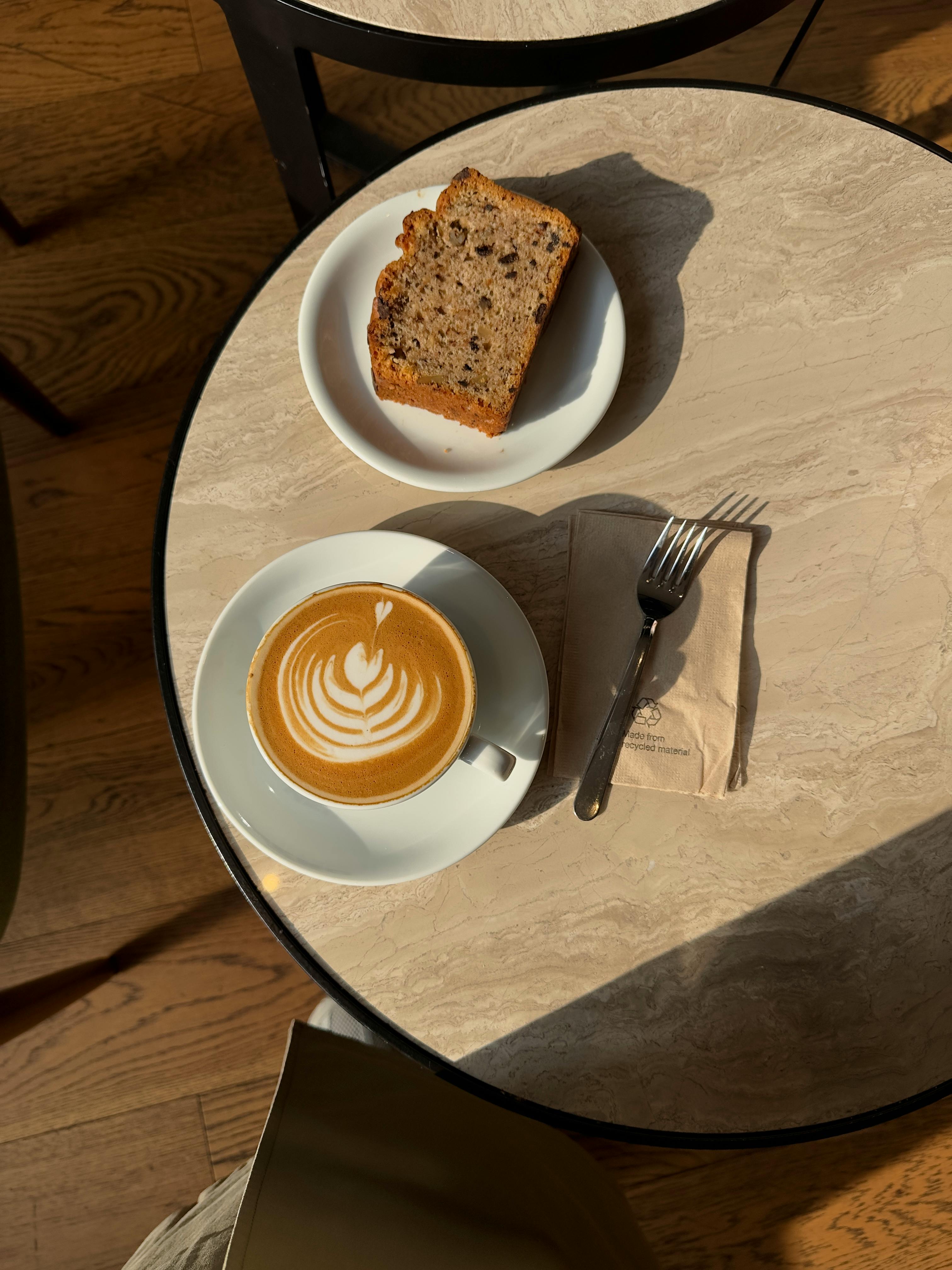 A cup of latte with intricate latte art paired with banana bread on a cafe table in sunlight.