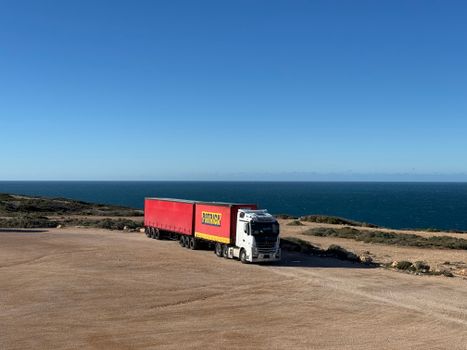 A white truck with a red trailer travels on a coastal road next to the ocean.